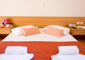 Habitación de hotel con una cama, dos almohadas, una colcha acolchada roja, dos toallas blancas, una mesita de noche con un libro, una cámara, una taza y un teléfono.