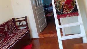 A room with a wooden floor, a bed with a red and beige patterned bedspread, a white chair, and a white ladder with a decorative cloth. - Traditional Camping ,Romania (Roșia Montană)