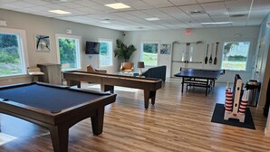 Une table de billard, une table de ping-pong et une table de jeu de fléchettes se trouvent dans une pièce bien éclairée, dotée d’un plancher en bois et de grandes fenêtres.