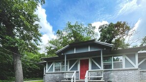 Une maison à deux étages avec une porte rouge, un porche à l’avant et un petit jardin devant, avec un arbre et un chemin en béton menant à la porte.