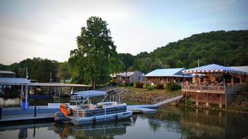 A lakeside dock with a boat, a restaurant with outdoor seating, and a forested hill in the background.