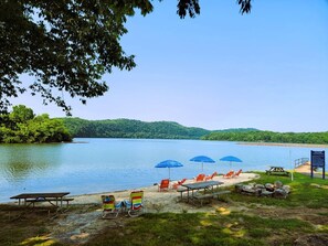 Una playa junto al lago con mesas de picnic, sillas de descanso y sombrillas.