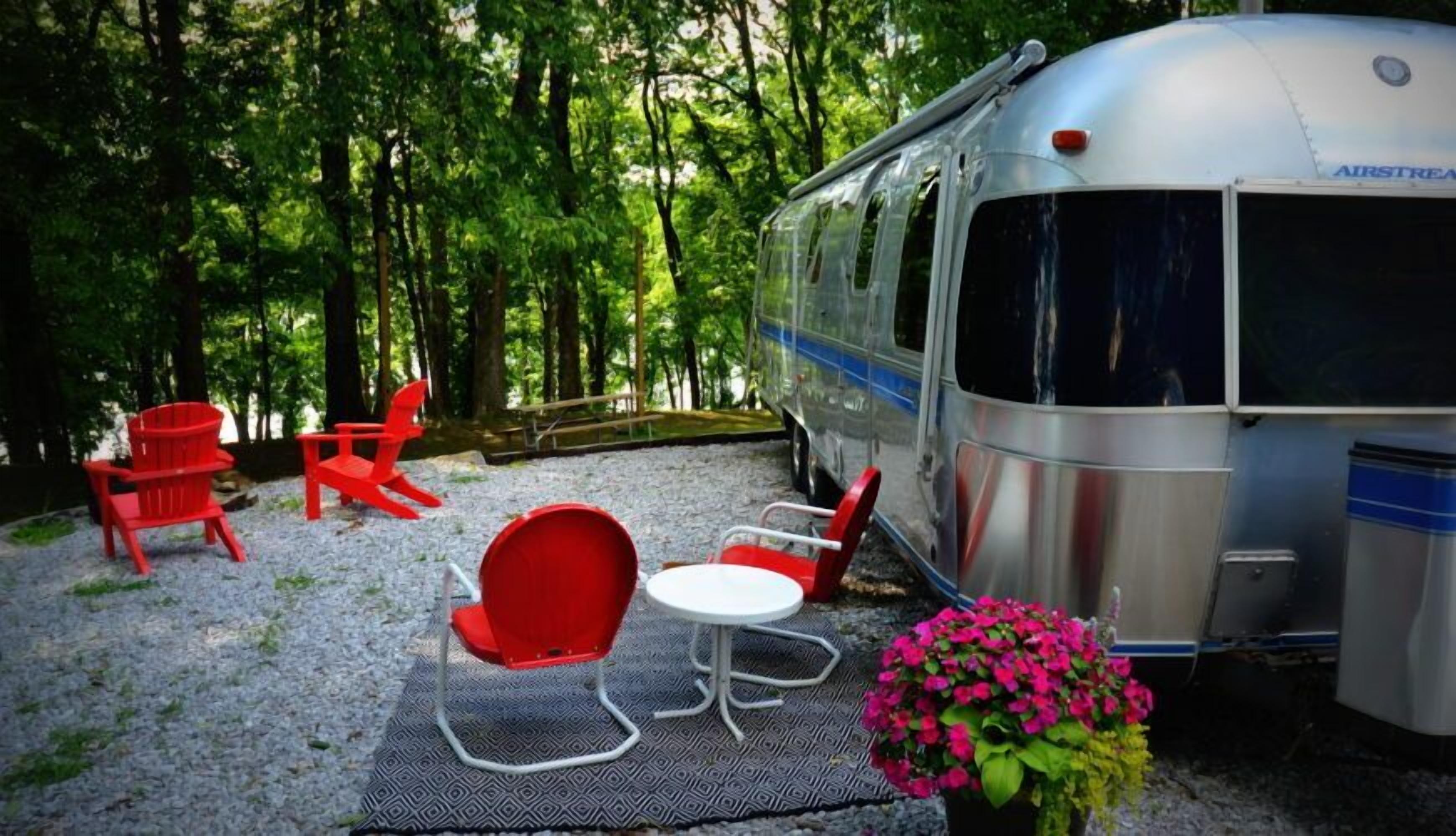 Une vieille caravane Airstream est garée dans une zone boisée, avec des chaises Adirondack rouges et une petite table.