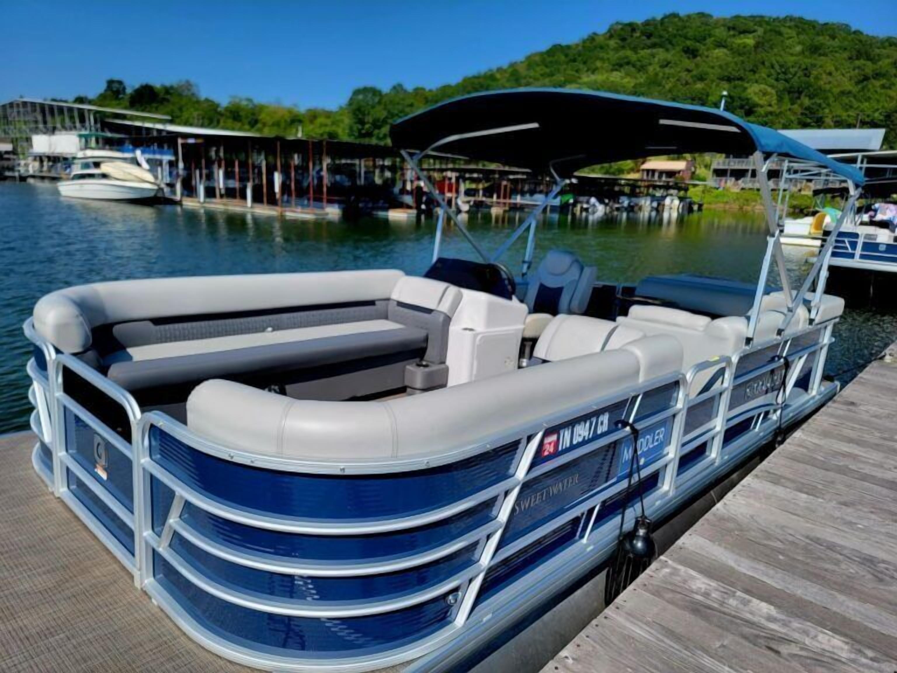 A pontoon boat docked at a pier with a covered seating area, a wakeboard, and a wakeboard tower.