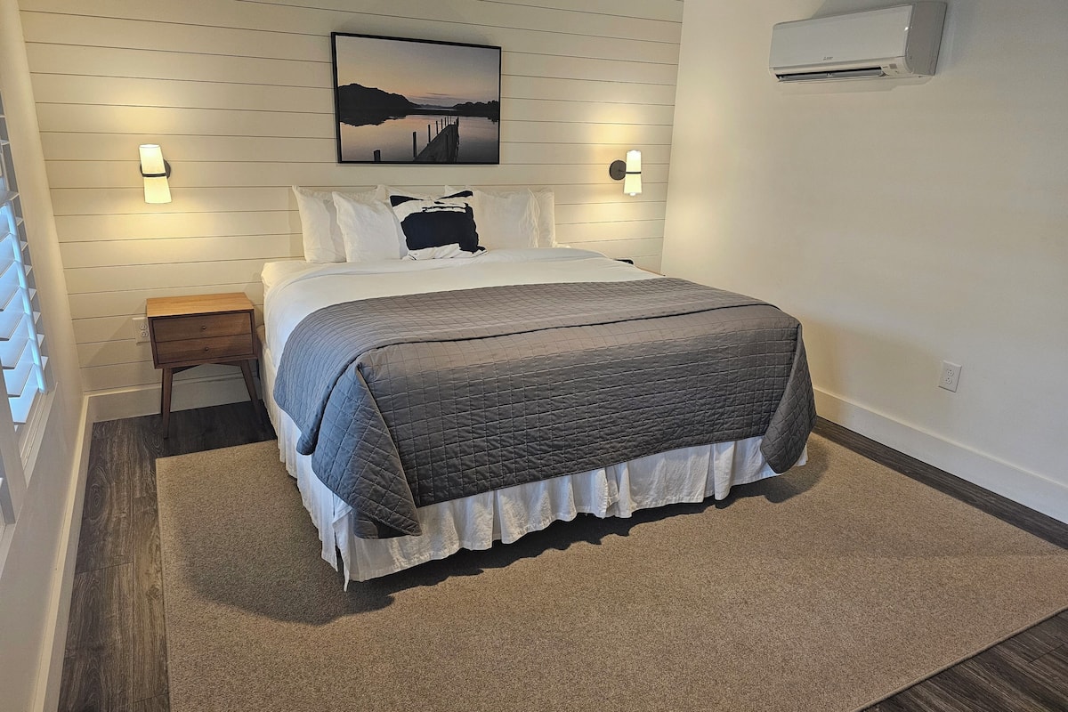 A neatly arranged bedroom with a bed, bedside tables, a wall-mounted air conditioner, and a framed picture above the bed.