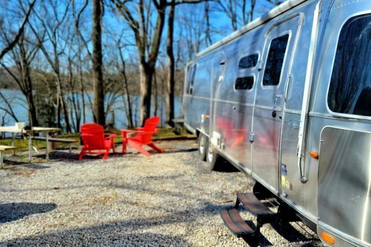 A silver RV parked on gravel with red chairs and a picnic table nearby.