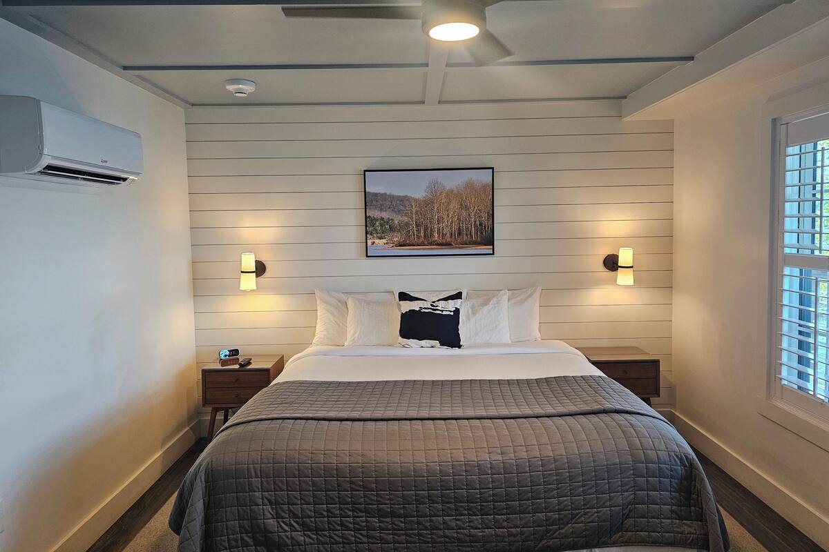 A modern bedroom with a bed, a ceiling fan, recessed lighting, a wall-mounted air conditioner, and a window with blinds.