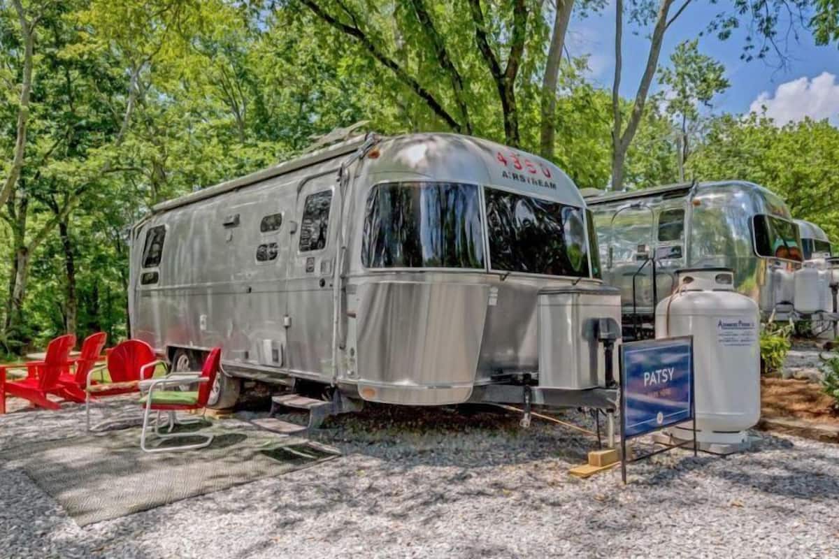 A silver RV with a large window, red chairs, and a water tank labeled "PATSY" in a wooded area.