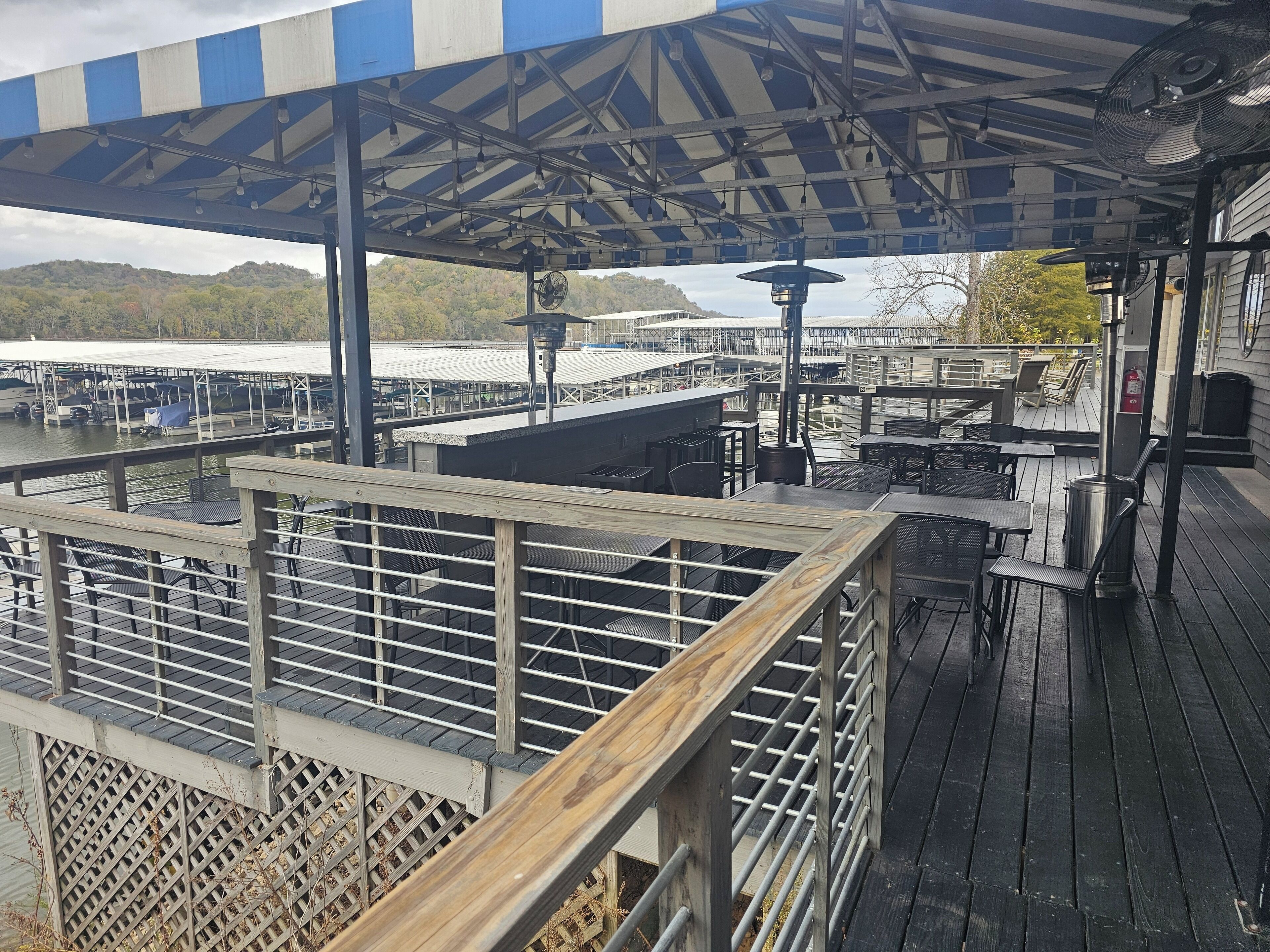 A covered outdoor seating area with tables and chairs, overlooking a body of water and docked boats.
