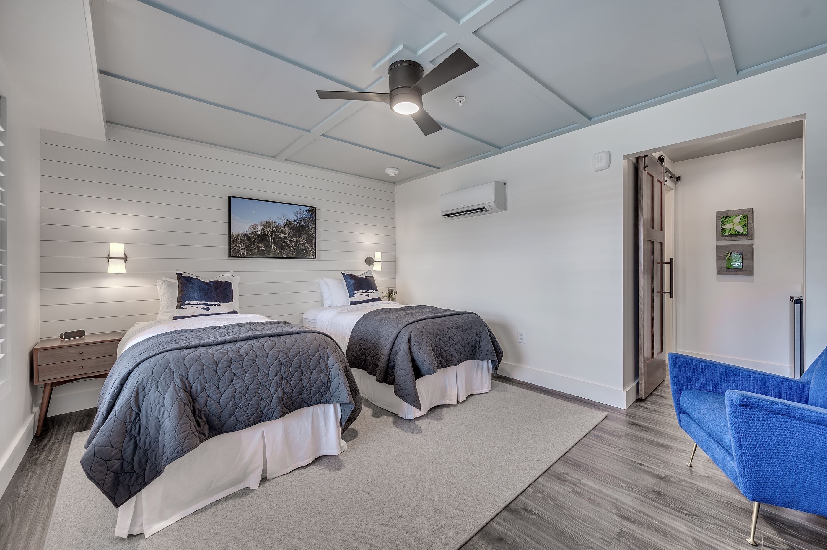 Un dormitorio con dos camas, un ventilador de techo, una mesita de noche, una silla azul y una puerta que da a otra habitación.
