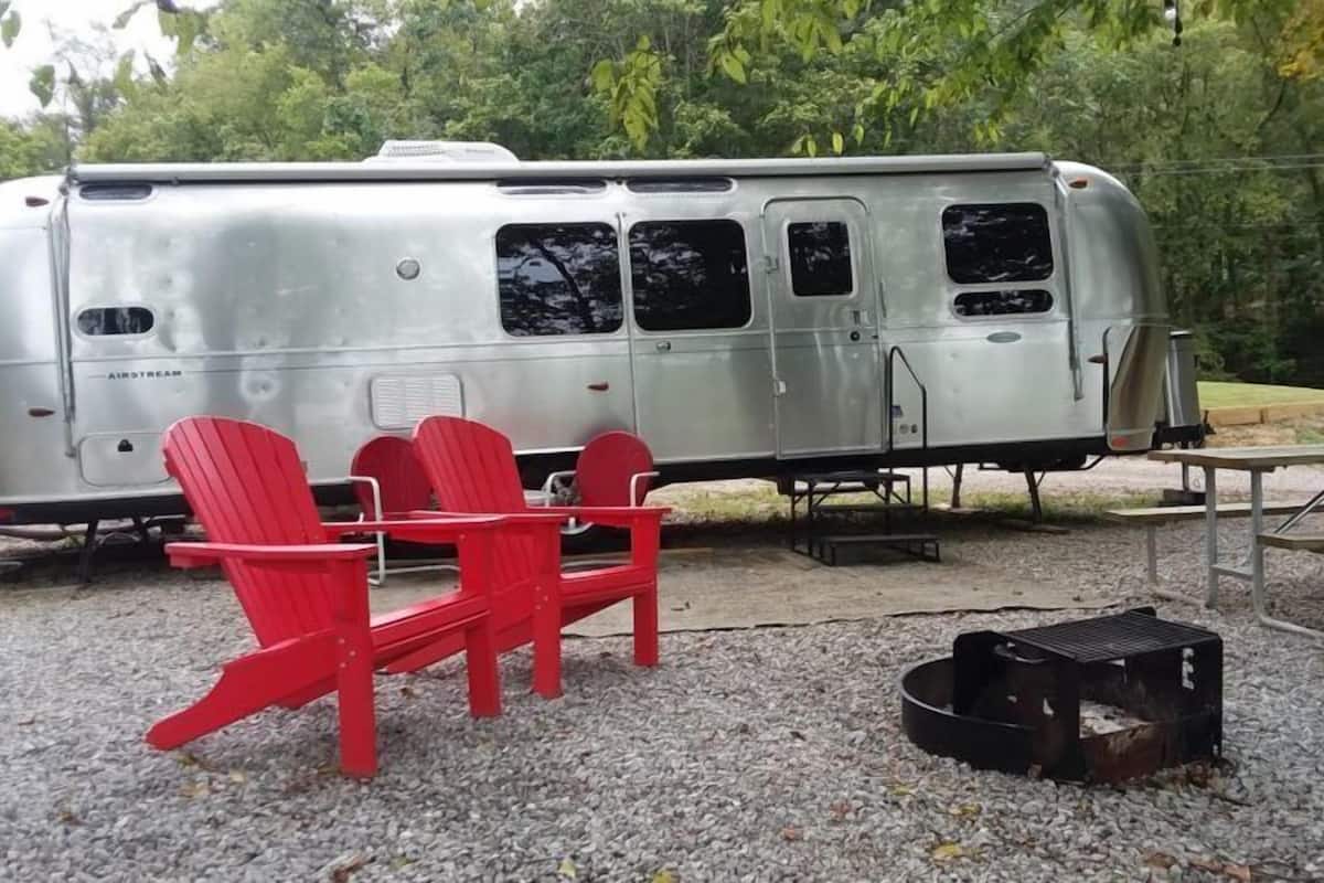 A silver RV with a kitchenette, two red Adirondack chairs, and a portable grill.