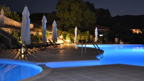 A nighttime swimming pool area with lounge chairs and umbrellas, surrounded by a stone wall and trees. - Villa Molyvos Castle (Lesvos)