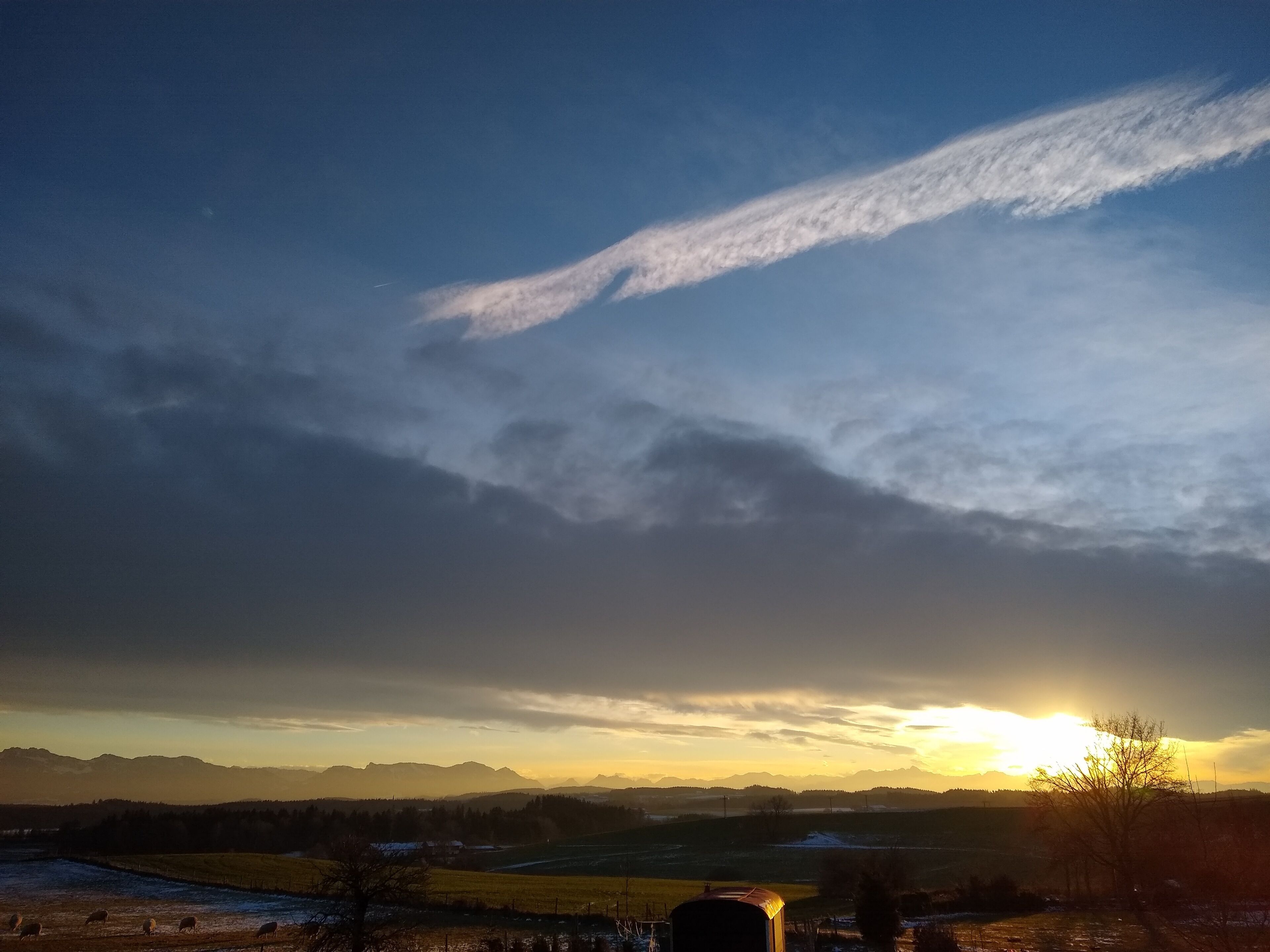 Ländliche Landschaft mit Sonnenuntergang, Bergen im Hintergrund und ein paar weidenden Schafen.