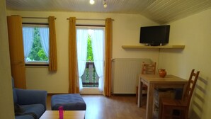 Una habitación con sofá, una mesita con una botella rosa, una silla de madera, una mesa de comedor con un jarrón y un televisor en la pared.