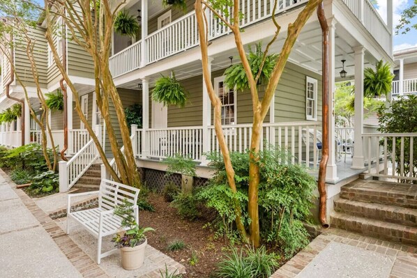 Un immeuble à plusieurs étages doté d’un porche couvert, de balustrades blanches et d’un petit jardin agrémenté d’un banc et de plantes en pot.