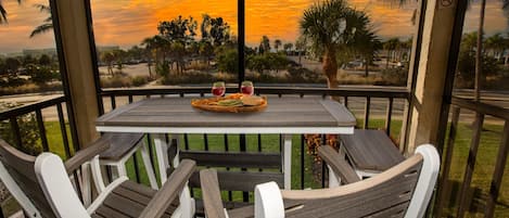 A wooden table with a woven basket and a glass of red drink on a balcony with a view of a sunset and palm trees.