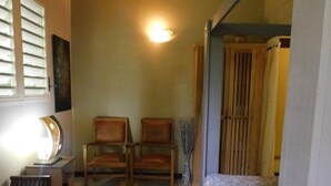 A small, cozy room with two wooden chairs, a bed with a patterned bedspread, a desk with a lamp, and a wall-mounted light fixture. - SOUS LES COCOTIERS STUDIO 2 CLIMATISE (Sainte-Rose)