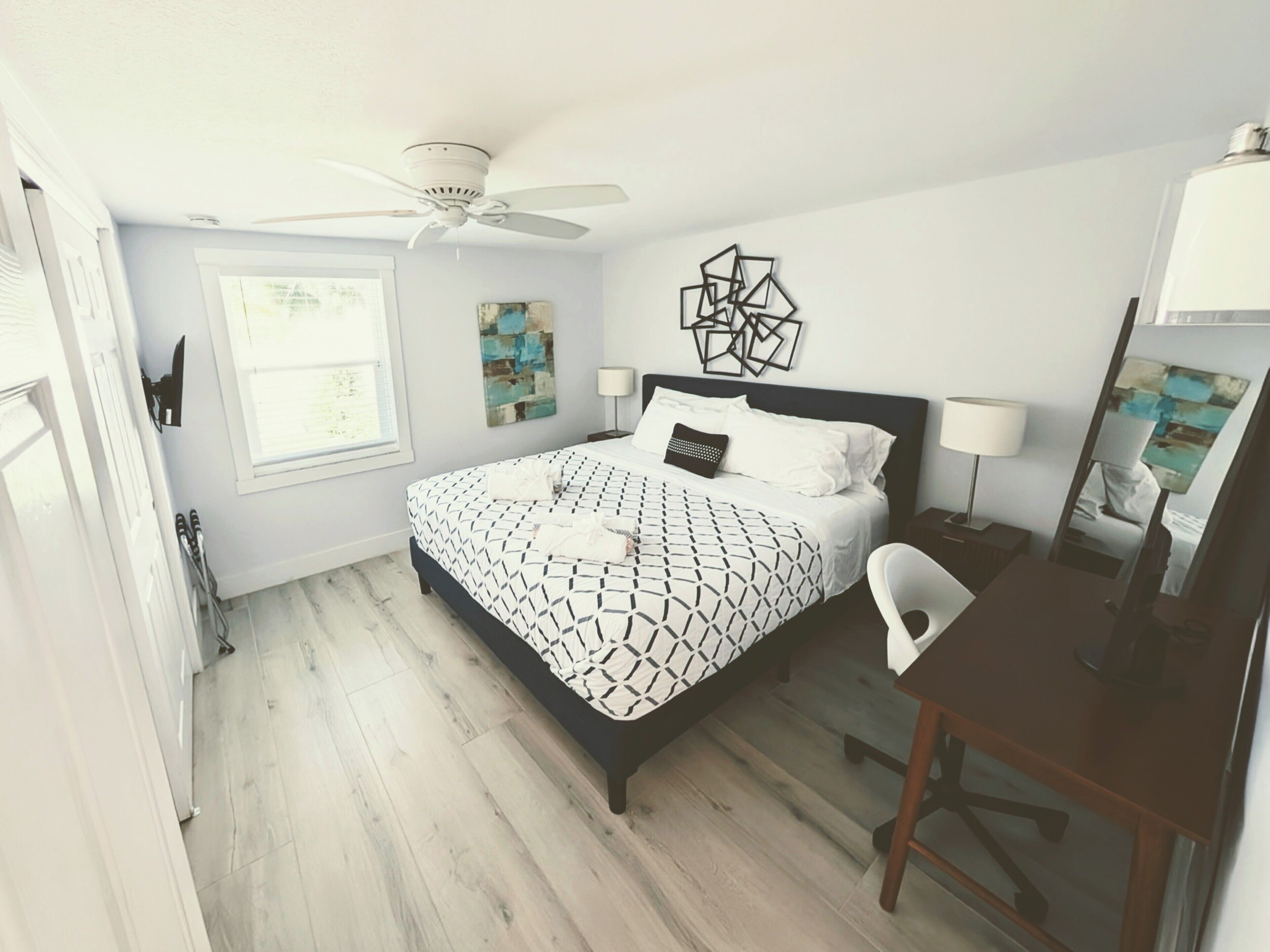 Un dormitorio con cama, escritorio, silla, ventilador de techo y obras de arte en las paredes.