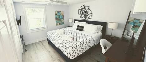 Un dormitorio con cama, escritorio, silla, ventilador de techo y obras de arte en las paredes.