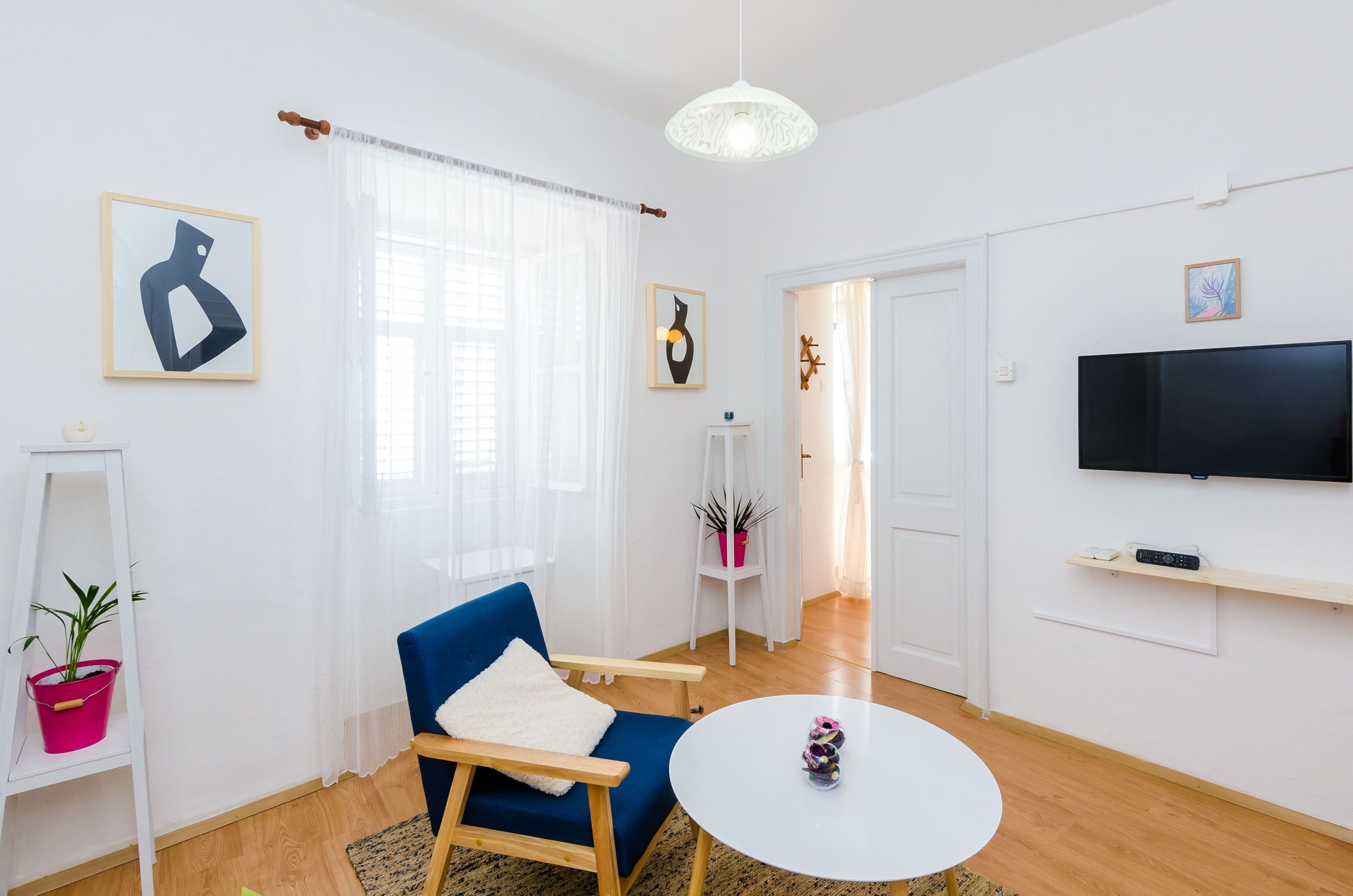 A modern living room with a blue armchair, a white coffee table, a flat-screen TV, and a large window with sheer curtains.
