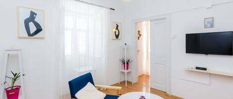 A modern living room with a blue armchair, a white coffee table, a flat-screen TV, and a large window with sheer curtains.