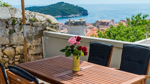 Un tavolo in legno con un vaso di fiori, un muro in pietra e vista sul mare e un'isola in lontananza.