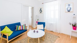 Un soggiorno con un divano blu, un tavolo rotondo bianco e una poltrona blu. Si nota una porta bianca, uno scaffale bianco con una pianta e una parete bianca con un quadro incorniciato.