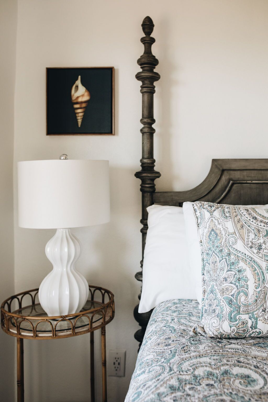 A bed with a patterned pillow, a white lamp on a round table, and a framed picture on the wall.