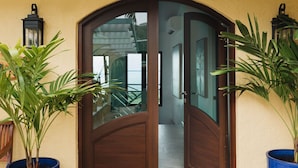 Una porta in legno con pannelli di vetro, affiancata da piante in vaso, che dà su un corridoio. Un tappetino d'ingresso con il logo di un cavalluccio marino è posto all'ingresso.