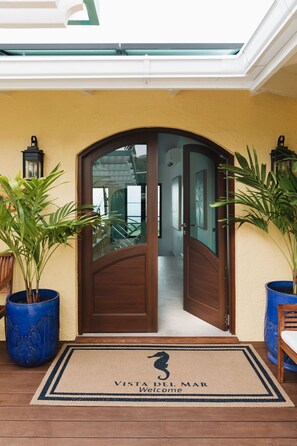 A wooden door with glass panels, flanked by potted plants, leading to a hallway. A welcome mat with a seahorse logo is placed at the entrance.