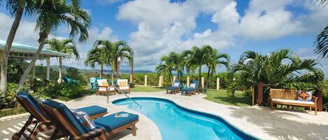 A swimming pool with lounge chairs and a wooden bench, surrounded by palm trees and a clear sky.