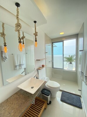 Une salle de bain moderne avec un grand miroir, un lavabo, des toilettes et une douche. Des serviettes sont accrochées au mur et, par la fenêtre, on peut admirer la vue sur l’océan.