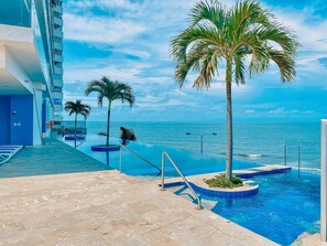 A pool area with a view of the ocean, palm trees, and a building with balconies.