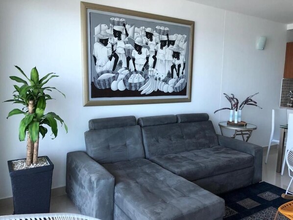 A living room with a grey sectional sofa, a framed black and white artwork, a potted plant, and a small round table with a vase.