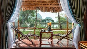 Una habitación con terraza de madera, dos sillones de descanso, una mesa con un jarrón de flores y vistas a un lago y árboles.