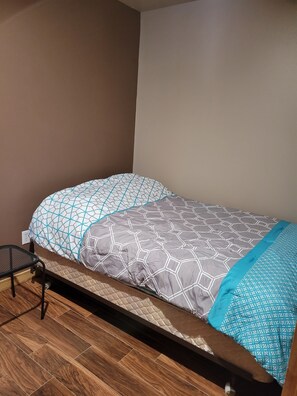 Un lit simple avec une couvre-lit et des oreillers à motifs géométriques, une table d’appoint en métal et un plancher en bois.