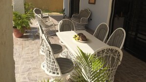Eine Terrasse im Freien mit einem weißen Tisch und Korbstühlen, umgeben von Blumentöpfen.