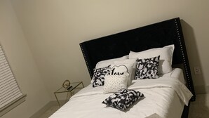 A neatly made bed with white bedding and black and white patterned pillows, a decorative wall piece above the bed, and a small bedside table with a lamp. - Apartment w/ Pool view 5 min from Galleria (Houston)