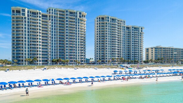 Un hotel frente a la playa con varios edificios altos, una playa arenosa con sombrillas azules y agua turquesa cristalina.