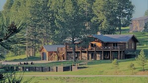 A log cabin with a porch and a small attached structure, surrounded by a forest and a field.