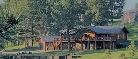 A log cabin with a porch and a small attached structure, surrounded by a forest and a field.