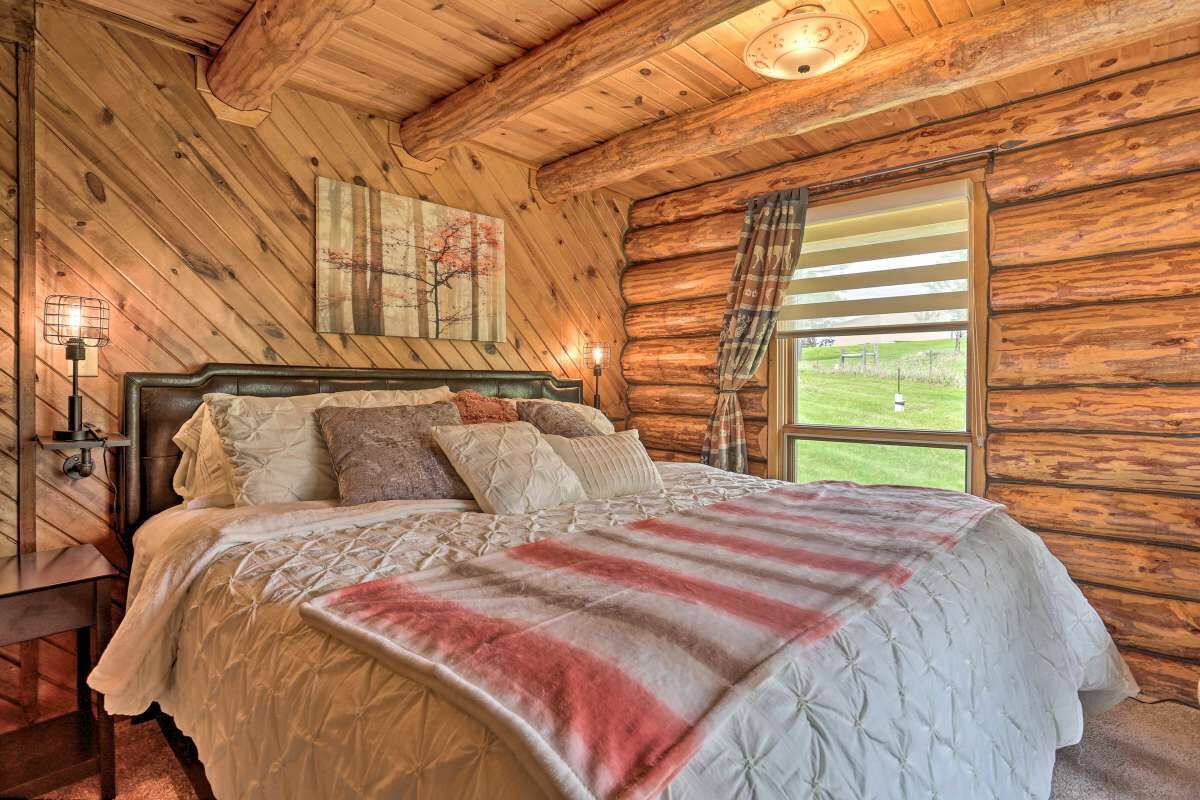 A log cabin bedroom with a large bed, a headboard, a painting on the wall, and a window with curtains.