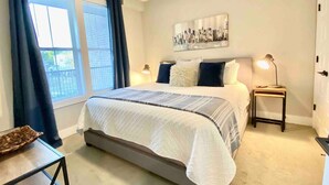 Une chambre à coucher avec un grand lit, une table de chevet avec une lampe, une fenêtre avec des rideaux et une œuvre d’art accrochée au mur.