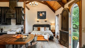A rustic bedroom with a wooden ceiling, a bed with floral bedding, a dining table with a meal, and a view of greenery outside. - Enchanted Oasis: Walk to Village, Pool, Hot Tub, Putting Green, 3 Houses (Pinehurst)