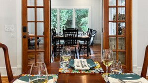 A dining area with a wooden table set for two, a view of the dining room through a glass door, and a view of the outdoors through another glass door. - Enchanted Oasis: Walk to Village, Pool, Hot Tub, Putting Green, 3 Houses (Pinehurst)