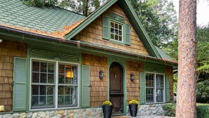 A rustic cabin with a stone foundation, green shutters, and a wooden front door. - Enchanted Oasis: Walk to Village, Pool, Hot Tub, Putting Green, 3 Houses (Pinehurst)