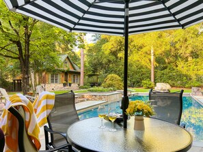 A poolside area with a round table, chairs, and an umbrella. - Enchanted Oasis | Walk to Village · Pool · Hot Tub · Putting Green · 3 Homes (Pinehurst)