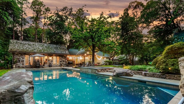 A pool area with a stone building, outdoor seating, and a view of trees. - Enchanted Oasis: Walk to Village, Pool, Hot Tub, Putting Green, 3 Houses (Pinehurst)