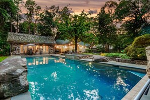 A pool area with a stone building, outdoor seating, and a view of trees. - Enchanted Oasis | Walk to Village · Pool · Hot Tub · Putting Green · 3 Homes (Pinehurst)
