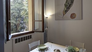 A dining area with a table set for two, a window with curtains, a radiator below the window, and a wall-mounted lamp.